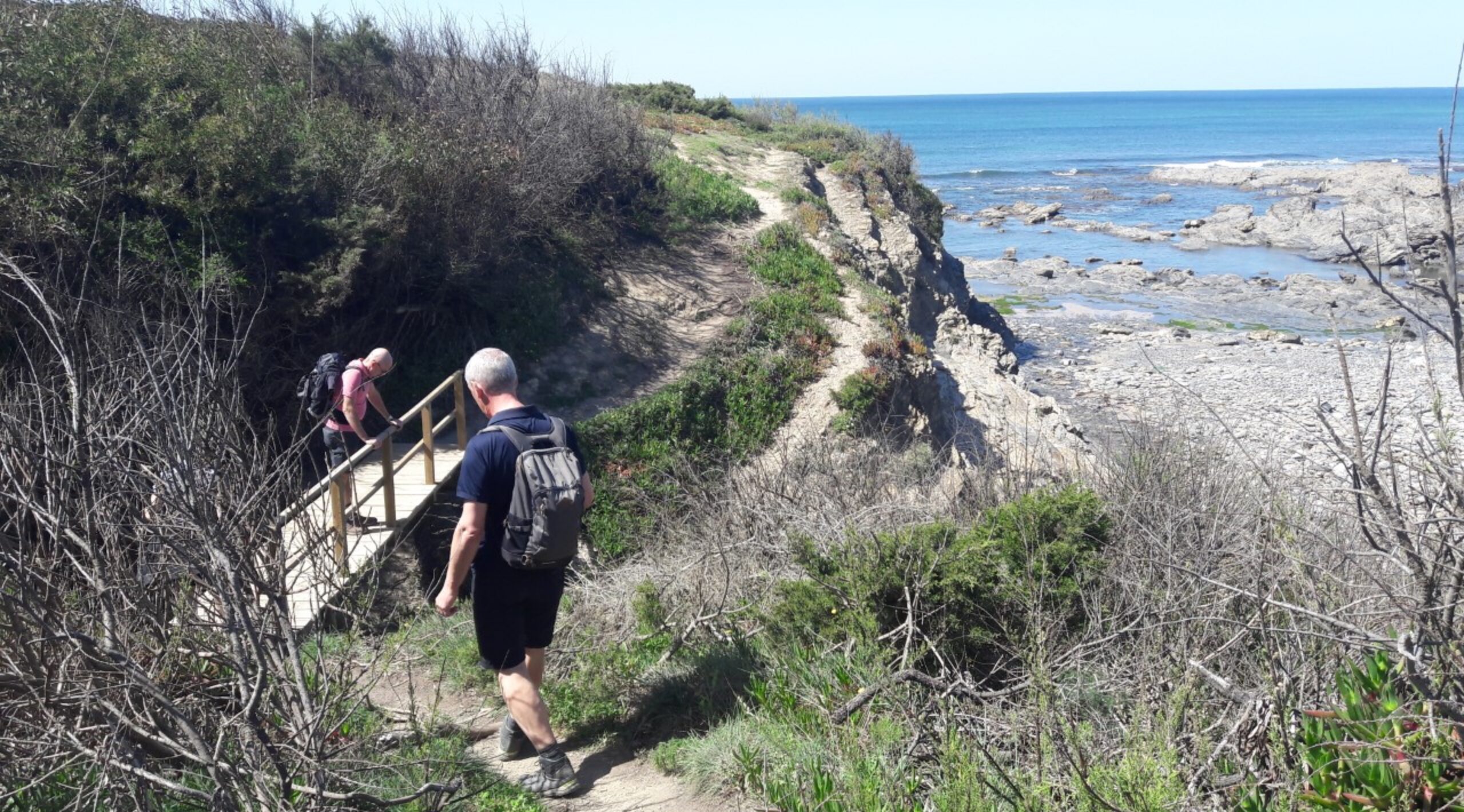 Twee wandelaars lopen over een houten promenade op een kustpad met uitzicht op een rotsachtig strand en een blauwe oceaan.