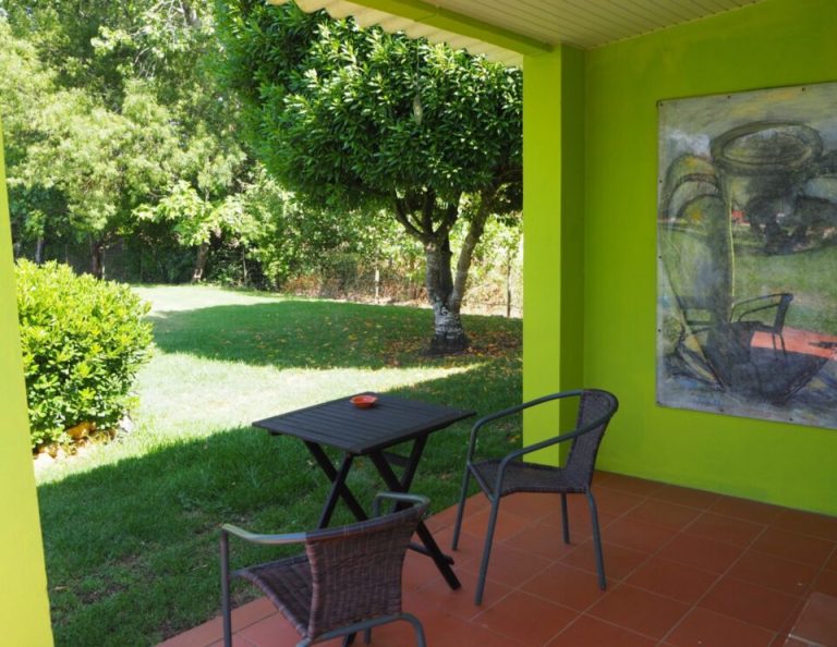 Eine helle lindgrüne überdachte Terrasse mit einem schwarzen Tisch und zwei Korbstühlen blickt auf einen üppigen Garten mit grünem Rasen, Bäumen und Hecken, an dessen Innenwand ein abstraktes Gemälde hängt.