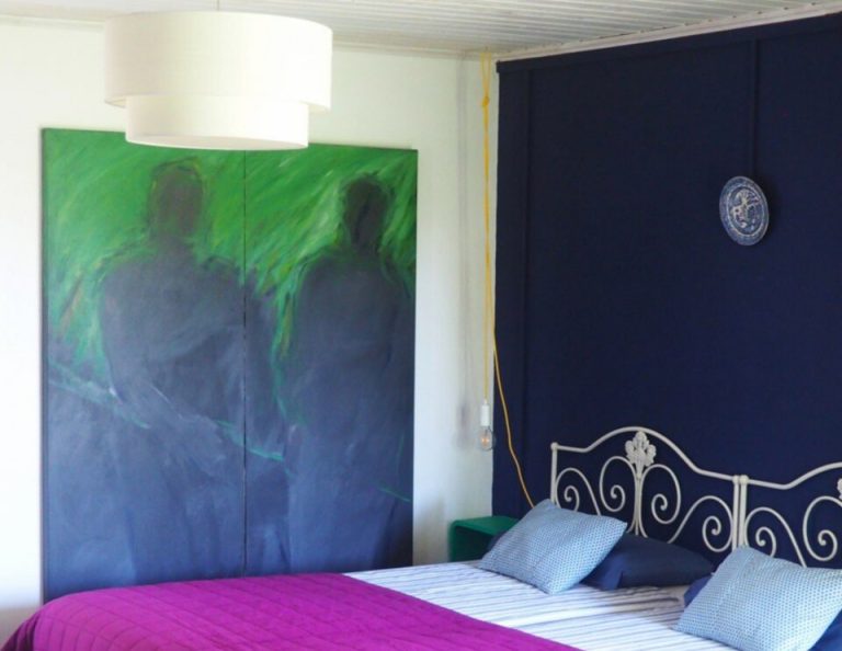 Ein modernes Schlafzimmer mit einem Bett mit einer leuchtend magentafarbenen Steppdecke und blau gestreiften Kissen, einem verschnörkelten Kopfteil aus weißem Metall, einem abstrakten Kunstwerk mit blau-grünem Farbverlauf über dem Bett und einer dunkeln marineblauen Akzentwand mit einer dekorativen Platte.