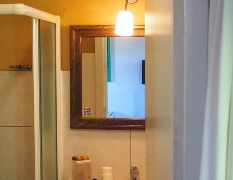 Ein moderner Waschtisch mit einem weißen, rechteckigen Waschbecken, einem holzgerahmten Spiegel mit Hängeleuchte darüber, einer Duschkabine auf der linken Seite und Toilettenartikeln auf dem Tresen.