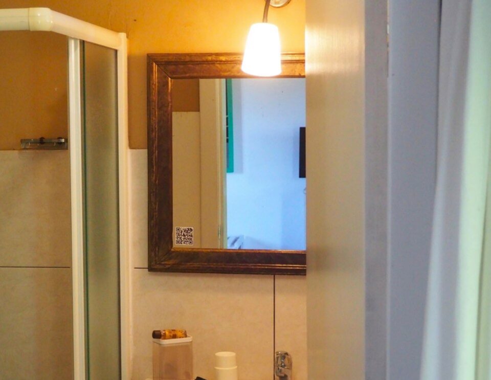 Ein moderner Waschtisch mit einem weißen, rechteckigen Waschbecken, einem holzgerahmten Spiegel mit Hängeleuchte darüber, einer Duschkabine auf der linken Seite und Toilettenartikeln auf dem Tresen.