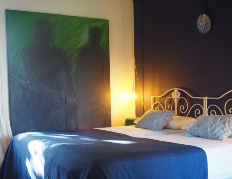 Ein modernes Schlafzimmer mit einem blauen Bett, einem verschnörkelten Kopfteil aus Messing, einem bunt gestreiften Teppich, einem großen abstrakten Kunstwerk in Grün und Blau, einer goldenen Akzentbeleuchtung und einer dekorativen Uhr an der dunklen Wand.