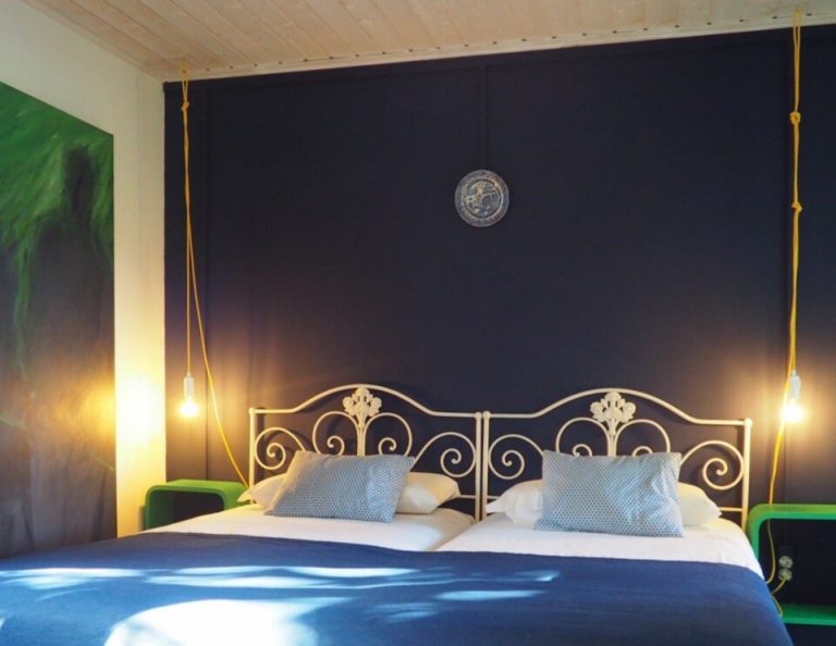 Ein modernes Schlafzimmer mit einem dekorativen goldenen Metallbettgestell vor einer dunklen marineblauen Wand, mit warmen Pendelleuchten auf beiden Seiten, weißen Kissen mit blau gemusterten Kissen, einem blauen Bettbezug, über den das Sonnenlicht fällt, einem grünen Kunstwerk an der linken Wand und einer dekorativen Platte über dem Bett.
