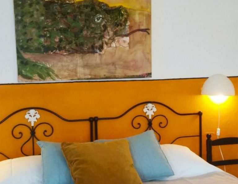 Ein helles Schlafzimmer mit einem verschnörkelten schwarzen Metallbettgestell vor einer leuchtend orangefarbenen Wand, dekoriert mit blauen und olivgrünen Kissen, einer weißen Lampe und einem abstrakten Landschaftsgemälde über dem Kopfteil.