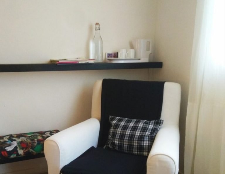 Ein weißer Sessel mit schwarz-weiß karierten Kissen und einer schwarzen Decke steht in einer Ecke neben einem Bett, darüber ein schwarzes, schwebendes Regal mit Dekorationsgegenständen und rechts ein Fenster mit durchsichtigen Vorhängen.