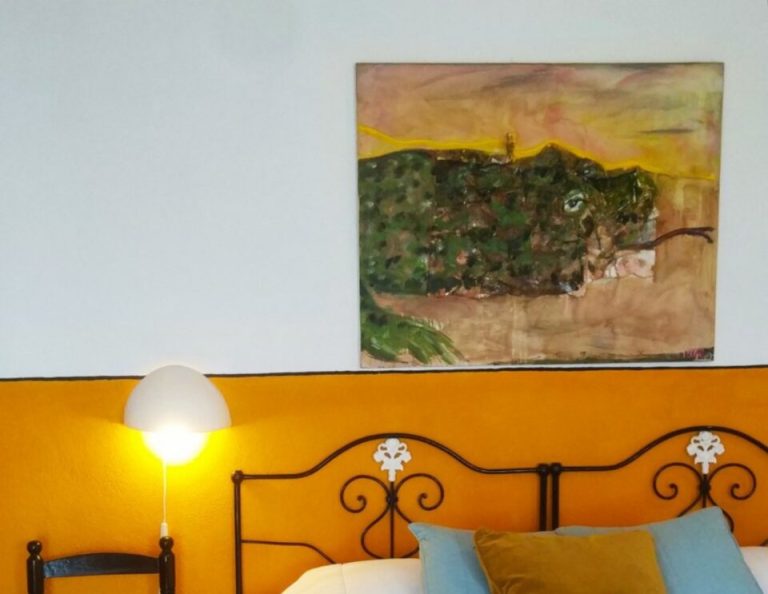 Ein helles Schlafzimmer mit einer leuchtend orangen Akzentwand verfügt über ein dekoratives schmiedeeisernes Kopfteil mit blauen und olivfarbenen Kissen, einen kleinen schwarzen Holzstuhl neben dem Bett, eine leuchtende Wandleuchte und ein Landschaftsgemälde über dem Kopfteil.