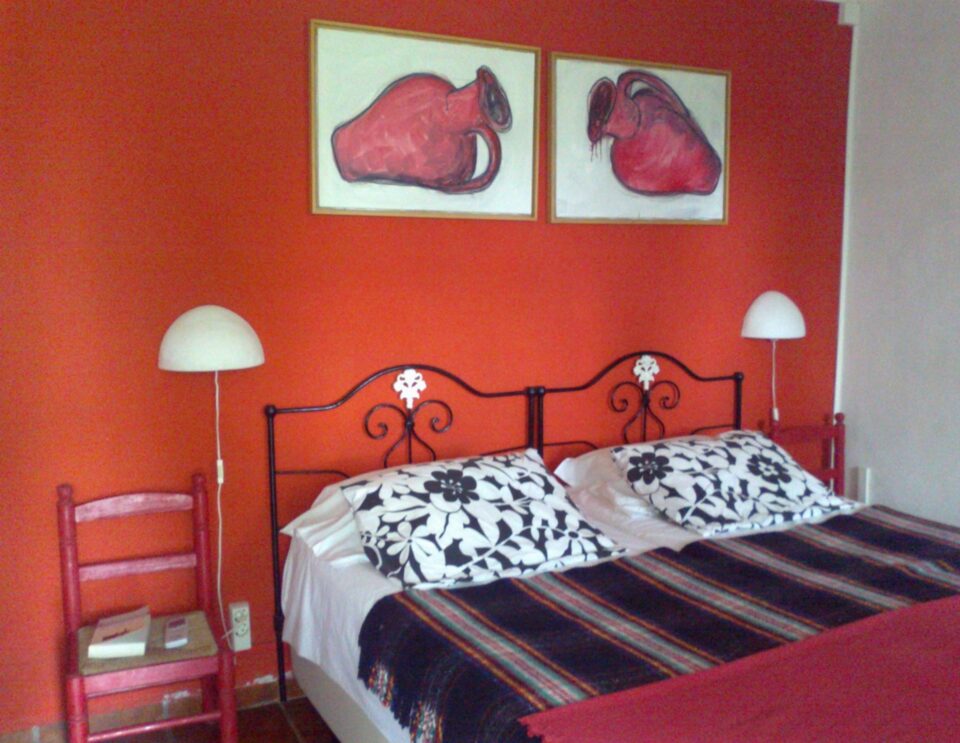 Ein leuchtend rotes Schlafzimmer mit einem dekorativen schwarzen Eisenbett mit weißen und schwarzen Blumenkissen, einer gestreiften Decke, zwei Kuppel-Pendelleuchten und zwei gerahmten roten anatomischen Herzzeichnungen an der Wand über dem Kopfteil.