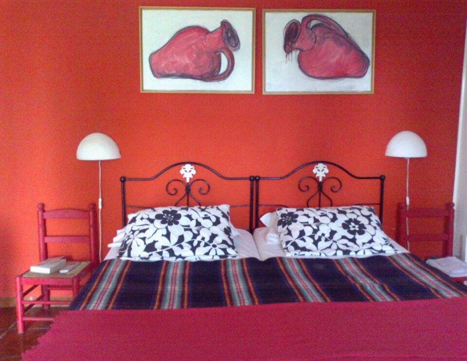 Ein leuchtend rotes Schlafzimmer mit zwei Einzelbetten mit verschnörkelten schwarzen Kopfteilen, schwarz-weiß gemusterten Kissen und gestreiften Decken, flankiert von roten Stühlen und weißen Kuppellampen, mit zwei rosa Aquarellbildern von Tieren an der roten Wand darüber.