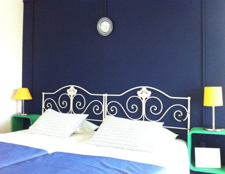 In einem modernen Schlafzimmer steht ein verziertes weißes schmiedeeisernes Bett vor einer dunkelblauen Wand, mit weißen Kissen und Bettzeug, flankiert von grünen Nachttischen mit gelben Tischlampen.