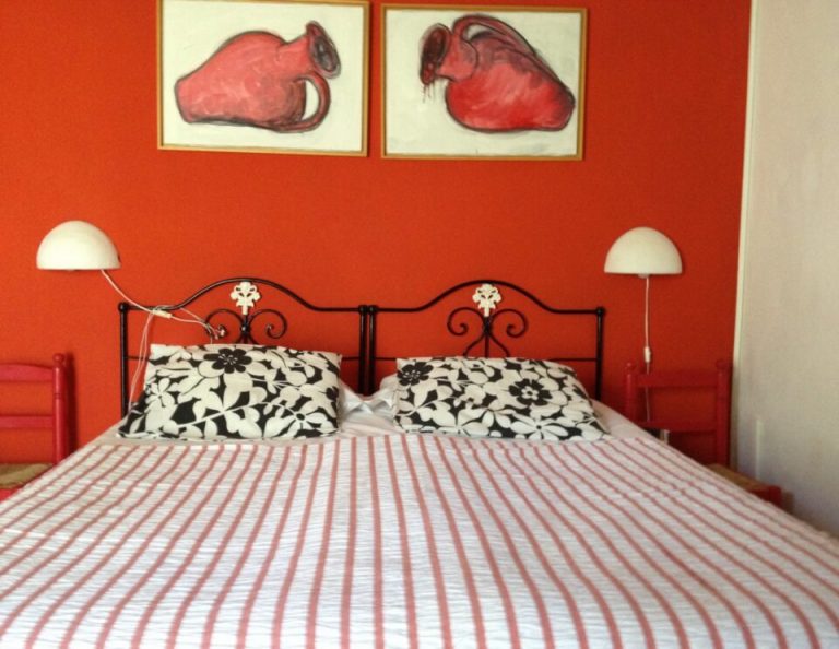 Ein leuchtend rotes Schlafzimmer mit einem schwarzen schmiedeeisernen Bettgestell, rot-weiß gestreifter Bettwäsche, schwarz-weiß gemusterten Kissen, weißen Kuppellampen auf beiden Seiten und zwei gerahmten roten anatomischen Herzzeichnungen, die über dem Kopfteil hängen.