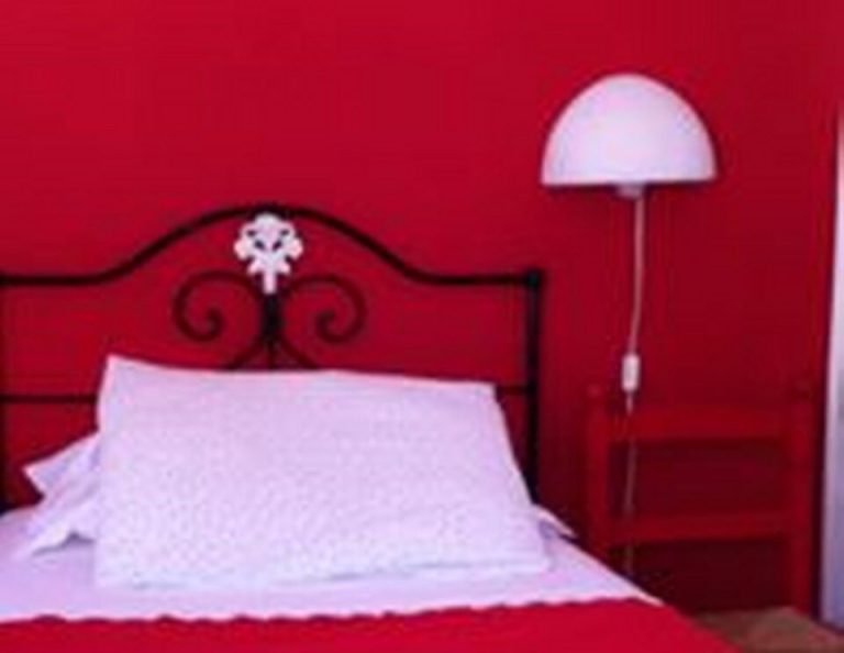 In einem Schlafzimmer mit tiefroten Wänden steht ein schwarzes Ornamentbett mit weißen Kissen und roter Bettwäsche, daneben eine weiße Stehlampe und eine Tür auf der rechten Seite.