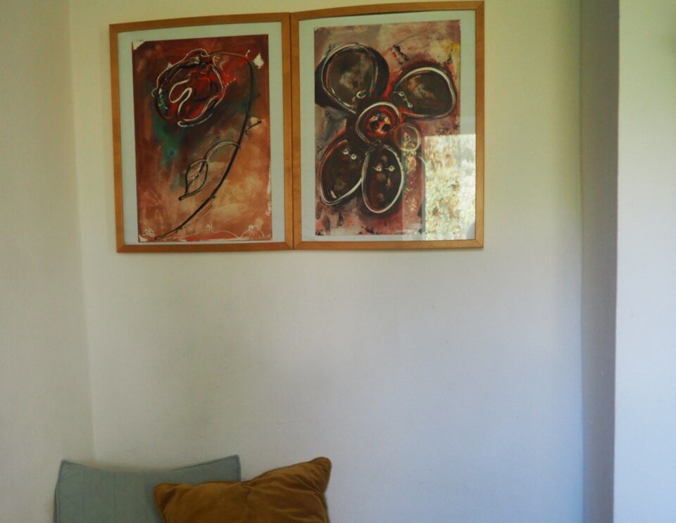 Eine minimalistische Schlafzimmerecke mit einem Bett mit senfgelbem Kissen und hellblauem Kissen vor einer weißen Wand, über dem zwei gerahmte abstrakte Kunstwerke in warmen Erdtönen hängen.