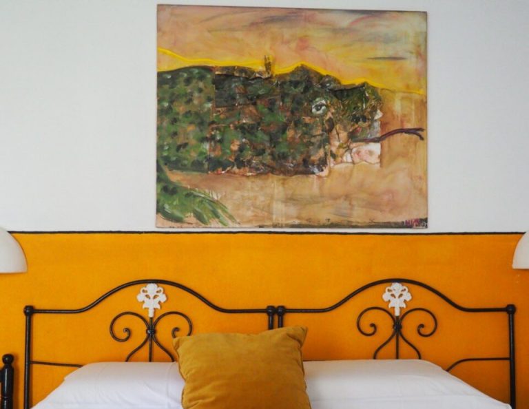 Ein Schlafzimmer mit einem leuchtend orangefarbenen Kopfteil und weißer Bettwäsche, mit einem dekorativen goldenen Kissen, einem verschnörkelten schmiedeeisernen Bettrahmen und einem darüber hängenden abstrakten Gemälde, das gedämpfte Erdtöne und dunkelgrüne Formen vor einem goldenen Himmel zeigt.