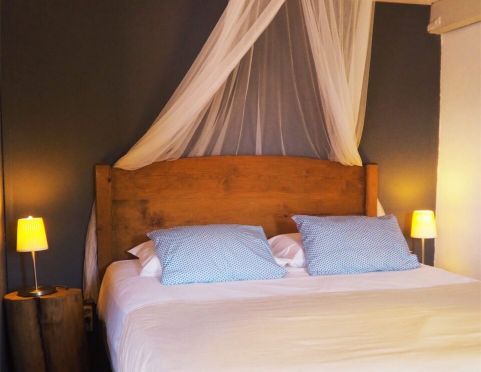 Ein Schlafzimmer mit einem hölzernen Himmelbett, das mit weißer Bettwäsche und blau gemusterten Kissen bezogen ist, flankiert von Nachttischen mit leuchtenden gelben Lampen vor warm getönten Wänden.