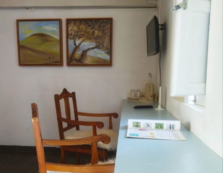 Ein minimalistischer Arbeitsbereich mit einem weißen Schreibtisch und Holzstühlen mit Schaffellkissen, zwei gerahmten Landschaftsbildern an der Wand, einem montierten Fernseher und einem Fenster mit weißen Jalousien.