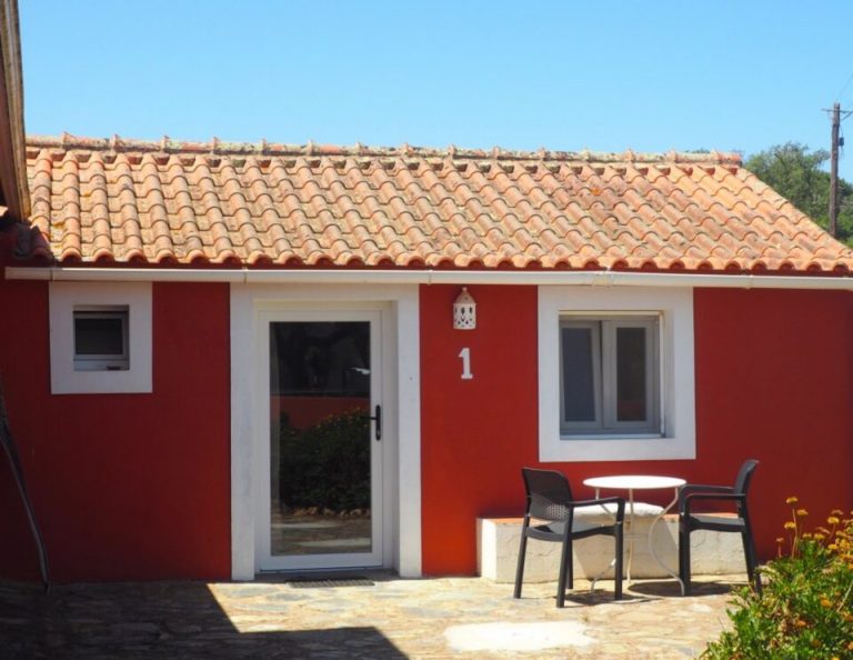 Ein leuchtend rotes Haus mit weißer Zierleiste und einem Dach aus Terrakotta-Ziegeln verfügt über eine Veranda mit zwei schwarzen Stühlen und einem runden Tisch sowie der Zahl "1" an der Eingangstür.