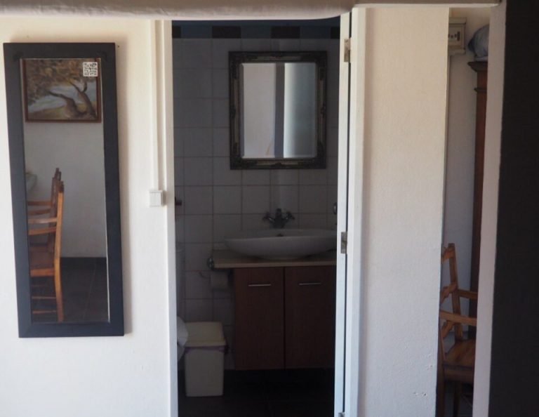 Eine Badezimmertür mit weißem Rahmen öffnet sich und gibt den Blick frei auf einen Waschtisch mit dunklem Schrank und schwarzen Fliesenwänden, mit einem gerahmten Spiegel über dem Waschbecken und einem kleinen Fenster.