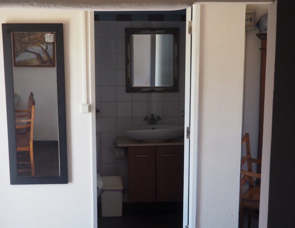 Eine Badezimmertür mit weißem Rahmen öffnet sich und gibt den Blick frei auf einen Waschtisch mit dunklem Schrank und schwarzen Fliesenwänden, mit einem gerahmten Spiegel über dem Waschbecken und einem kleinen Fenster.