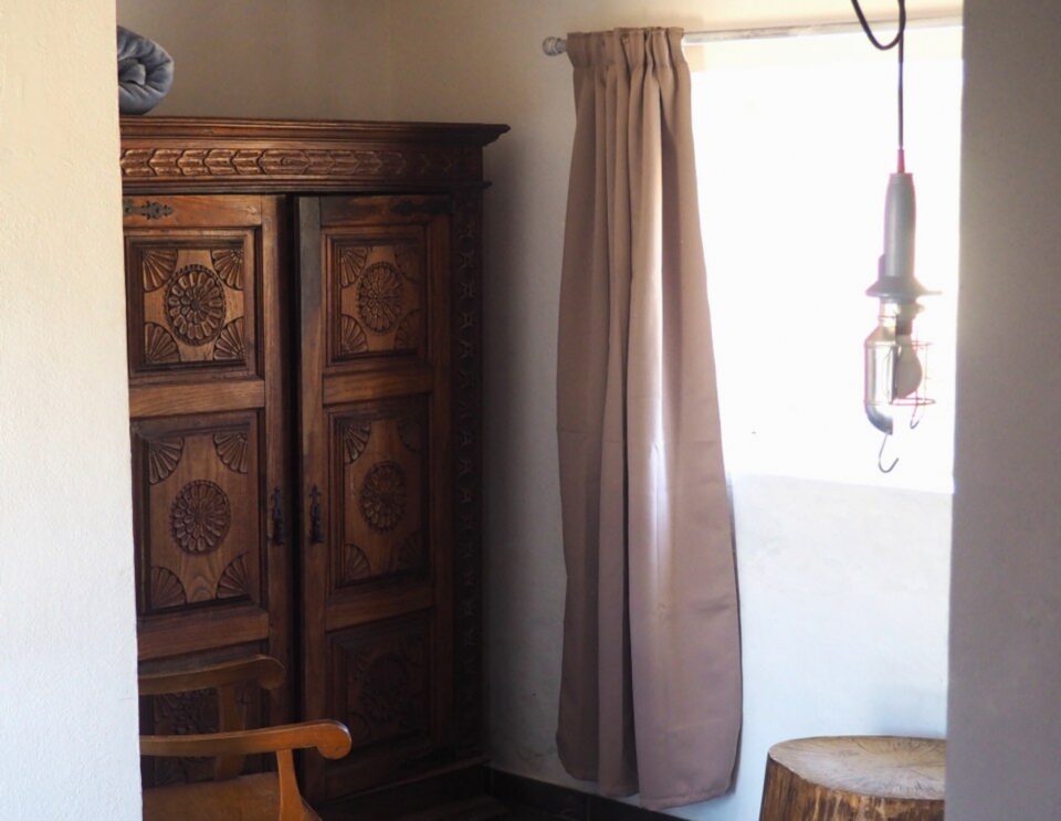 Eine rustikale Schlafzimmerecke mit einem kunstvoll geschnitzten Holzschrank, einem Holzstuhl, einem Beistelltisch aus Baumstümpfen und einem Fenster mit hellbraunen Vorhängen und einer Hängelaterne.