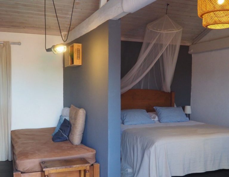 Ein modernes Schlafzimmer mit einem Himmelbett mit weißen Vorhängen, Holzmöbeln, darunter ein Sessel mit Kissen, Pendelleuchten und dunklen Fliesenböden.