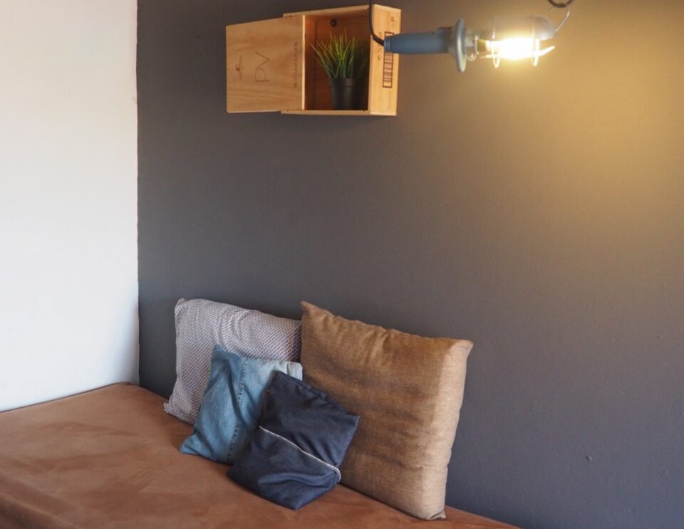 In einer minimalistischen Schlafzimmerecke steht ein Bett mit brauner Bettwäsche und dekorativen Kissen vor einer zweifarbigen grauen Wand, darüber ein hölzernes Wandregal mit einer Topfpflanze und rechts daneben eine Hängeleuchte.