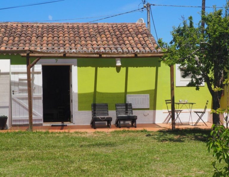 Ein helles, lindgrünes und weißes Häuschen mit einem Dach aus Terrakotta-Ziegeln verfügt über eine überdachte Terrasse mit schwarzen Liegestühlen und einem kleinen Tisch, von der aus man auf eine Wiese mit Bäumen blickt.