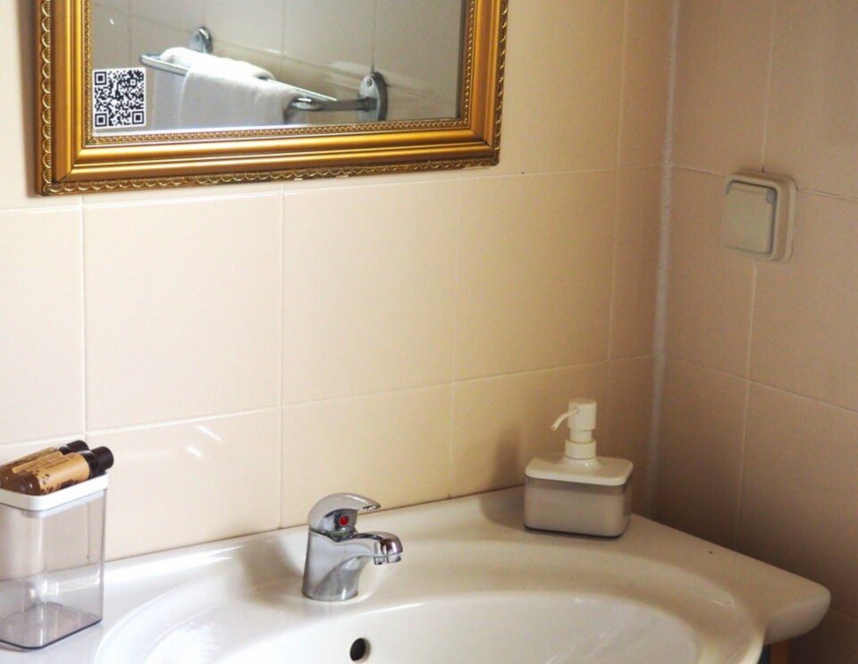 Ein Waschbecken mit verchromtem Wasserhahn und Seifenspender, ein gerahmter Spiegel mit Goldverzierung darüber, cremefarbene Fliesenwände und ein kleiner Behälter auf der linken Arbeitsplatte.
