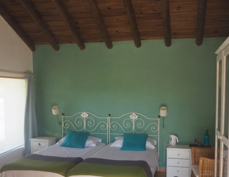 Ein helles Schlafzimmer mit mintgrünen Wänden und freiliegenden Holzbalken an der Decke verfügt über zwei Einzelbetten mit einem verschnörkelten Metallkopfteil, blaue und grüne Bettwäsche, blaugrüne Kissen und weiße Nachttische auf beiden Seiten.