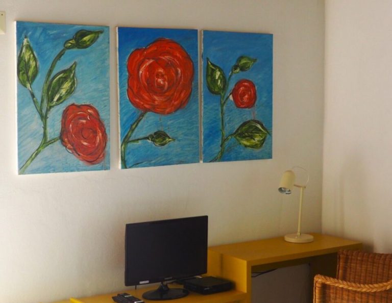 Drei Gemälde von roten Rosen mit grünen Knospen auf blauem Hintergrund hängen über einem hölzernen Schreibtisch mit Computermonitor, Lampe und Korbsessel.