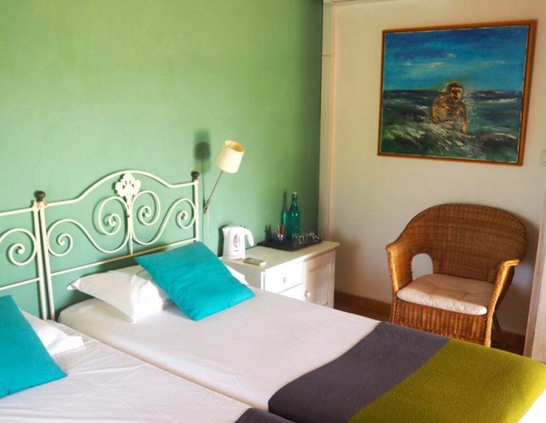 In einem hellen Schlafzimmer mit mintgrünen Wänden stehen ein weißes, verziertes Metallbett mit türkisfarbenen und weißen Kissen, ein Korbsessel, ein Nachttisch mit Lampe, ein gerahmtes Gemälde mit Meeresmotiven und eine farbenfrohe Decke in Gelb und Anthrazit.