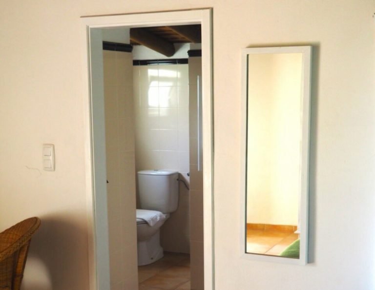 Innenansicht eines Schlafzimmers mit einer offenen Tür, die zu einem Badezimmer führt, in dem sich eine Toilette, rechts ein Wandspiegel und ein Holzfußboden befinden.