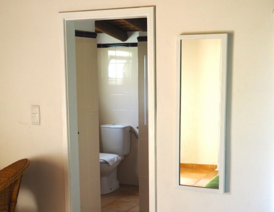 Innenansicht eines Schlafzimmers mit einer offenen Tür, die zu einem Badezimmer führt, in dem sich eine Toilette, rechts ein Wandspiegel und ein Holzfußboden befinden.