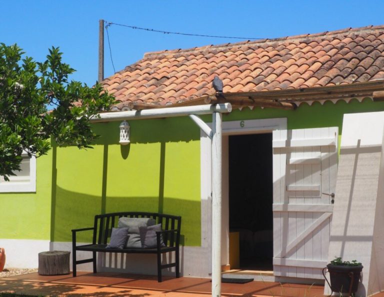 Ein helles, lindgrünes Haus mit einem Dach aus Terrakotta-Ziegeln verfügt über eine überdachte Terrasse mit einer schwarzen Bank, einer weißen Pergola und Topfpflanzen, die einen Blick auf einen grasbewachsenen Rasen unter blauem Himmel bietet.