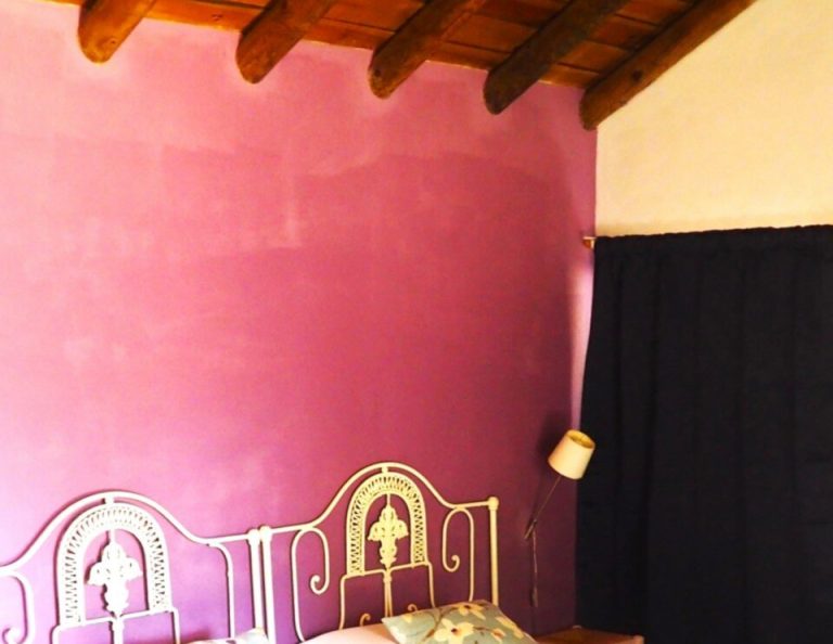 Ein Schlafzimmer mit leuchtend rosafarbenen Wänden, freiliegenden Holzbalken an der Decke, einem verschnörkelten Metallbettgestell in Gold und Weiß, weißen Kissen und einem schwarzen Vorhang an der rechten Wand.