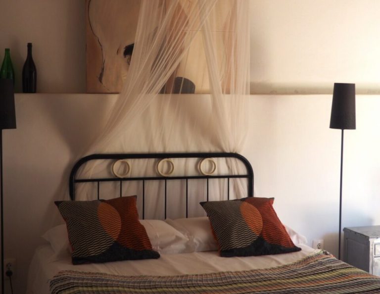 Ein gemütliches Schlafzimmer mit einem schwarzen Metallbettgestell mit dekorativen Kissen, einem weißen Baldachin, einer strukturierten Hängeleuchte darüber, einer hohen Stehlampe und Weinflaschen auf einem Regal.