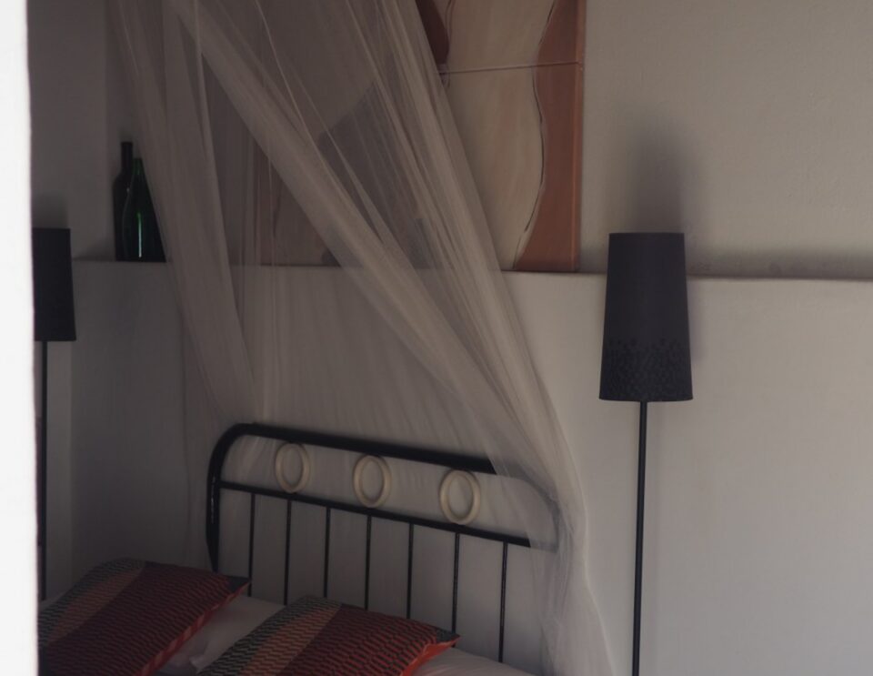 Ein Schlafzimmer mit einem schwarzen Metallbettgestell und weißen Vorhängen, dekoriert mit rot und schwarz gemusterten Kissen, einer Stehlampe mit schwarzem Schirm und einem Kunstwerk an der Wand über dem Bett.