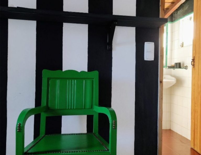 Ein hellgrüner Holzstuhl steht vor einer schwarz-weiß gestreiften Wand, und durch eine offene Tür auf der rechten Seite ist ein Badezimmer zu sehen.