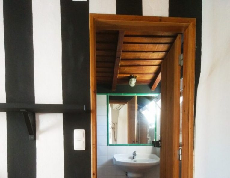 Eine Badezimmertür mit einem Waschbecken, einem Spiegel mit grünem Rahmen und einer Holzdecke, die hinter der offenen Holztür zu sehen ist, von einem Raum mit schwarz-weiß gestreiften Wänden und Holzfußboden aus gesehen.