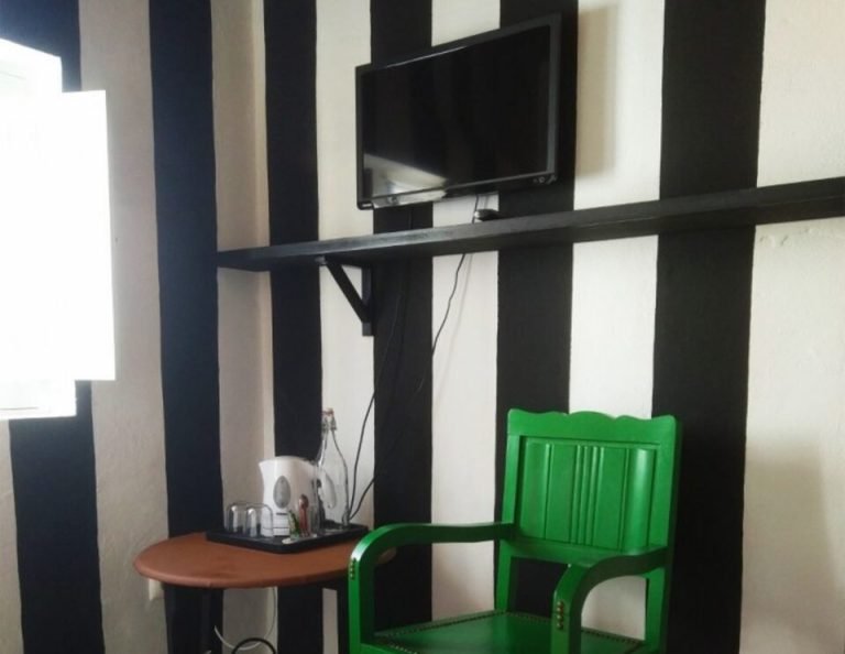 Ein leuchtend grüner Holzstuhl steht vor einem kleinen runden Tisch, über dem ein Fernseher an der Wand montiert ist, vor einer schwarz-weiß gestreiften Tapete in einem minimalistischen Raum.