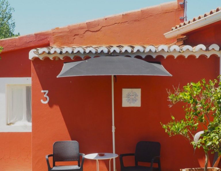 Ein leuchtend orange-rotes Gebäude mit weißer Dachverzierung verfügt über einen schattigen Sitzbereich mit zwei schwarzen Stühlen, einem kleinen Tisch und einem grauen Sonnenschirm, in dessen Nähe sich Topfpflanzen befinden.