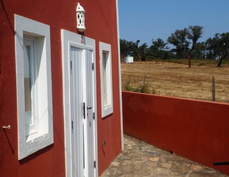 Ein rotes Gebäude mit weißen Türen und Fenstern steht in einer kargen Landschaft mit vereinzelten Bäumen und trockenem Grasland unter einem klaren blauen Himmel.