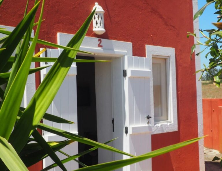 Ein kleines rot-weißes Gebäude mit der Nummer 2, mit einer weißen Tür und einem weißen Fenster, mit grünen Pflanzenblättern im Vordergrund und einer weißen dekorativen Laterne über dem Eingang.