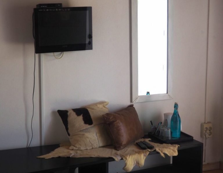 Ein minimalistisches Zimmer mit einer schwarzen Sitzbank vor einer weißen Wand, darauf hellbraune und braune Kissen, eine blaue Flasche und eine Fernbedienung, darüber ein an der Wand montierter Fernseher und ein hohes schmales Fenster auf der rechten Seite.