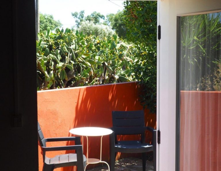 Auf einer hellen Terrasse mit orange-roter Wand stehen zwei schwarze Stühle, die einen kleinen weißen runden Tisch flankieren, mit Blick auf einen üppigen Garten mit grünen Pflanzen und Bäumen.