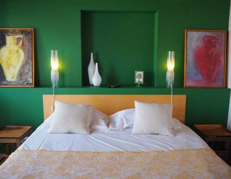 Ein modernes Schlafzimmer mit grüner Akzentwand verfügt über ein gelb-orangefarbenes Kopfteil, weiße Bettwäsche mit gemusterten Kissen, beleuchtete Wandleuchter an beiden Seiten und gerahmte Kunstwerke mit klassischen Gefäßen über dem Bett.