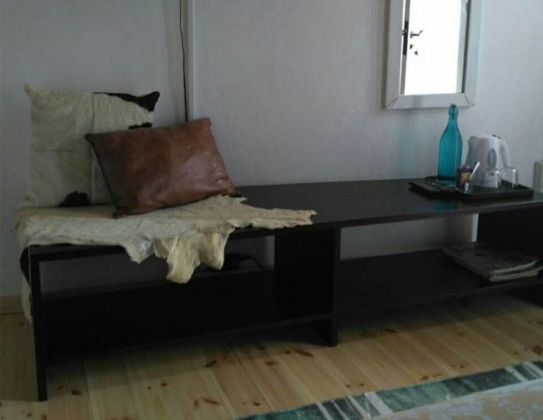Ein minimalistisches Schlafzimmer mit einem schwarzen Bett mit cremefarbenen und braunen Kissen, einem schwarzen Schreibtisch, der aus dem Bettrahmen herausragt, einem Spiegel über dem Schreibtisch, einer türkisfarbenen Flasche und einem weißen Gerät auf der Schreibtischoberfläche sowie einem hellen Holzfußboden.