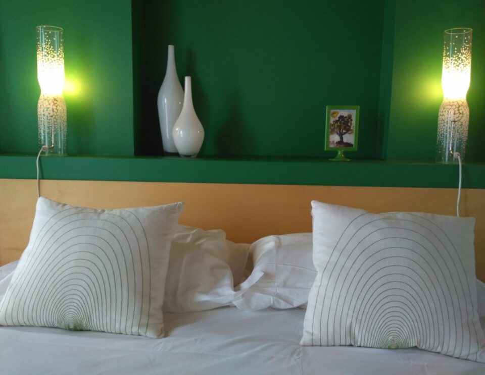 Ein modernes Schlafzimmer mit einem Kopfteil aus Naturholz und weißer Bettwäsche verfügt über zwei dekorative Kissen mit konzentrischen Kreismustern, flankiert von beleuchteten zylindrischen Lampen an einer grün- und hellbraunen Akzentwand.