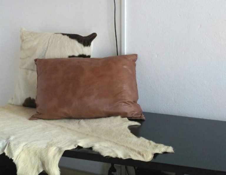 Zwei dekorative Kissen, gestapelt an einer weißen Wand, mit einem cremefarbenen Kissen hinter einem braunen Lederkissen, neben einem schwarzen Tisch mit einer cremefarbenen Felldecke.
