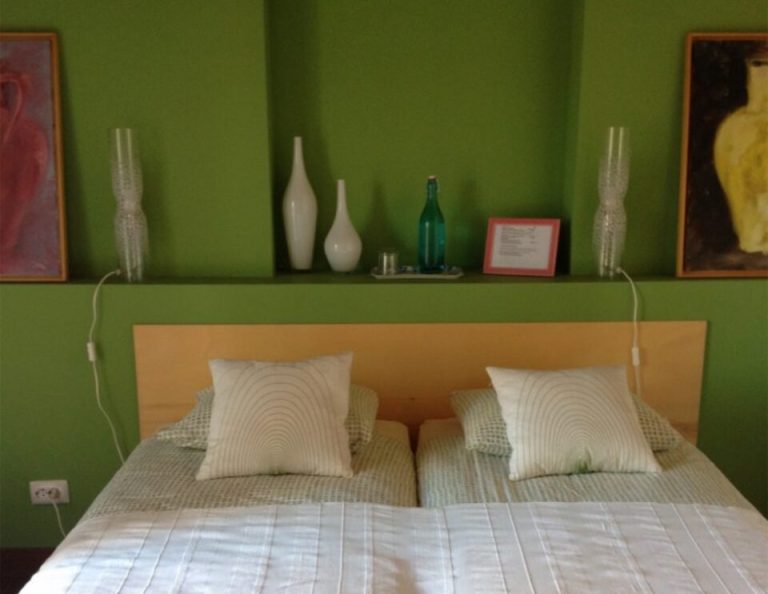 Ein modernes Schlafzimmer mit einem hölzernen Kopfteil und weiß gestreifter Bettwäsche vor einer hellgrünen Wand, in die ein Regal mit dekorativen Flaschen, Vasen und gerahmten Kunstwerken eingelassen ist.