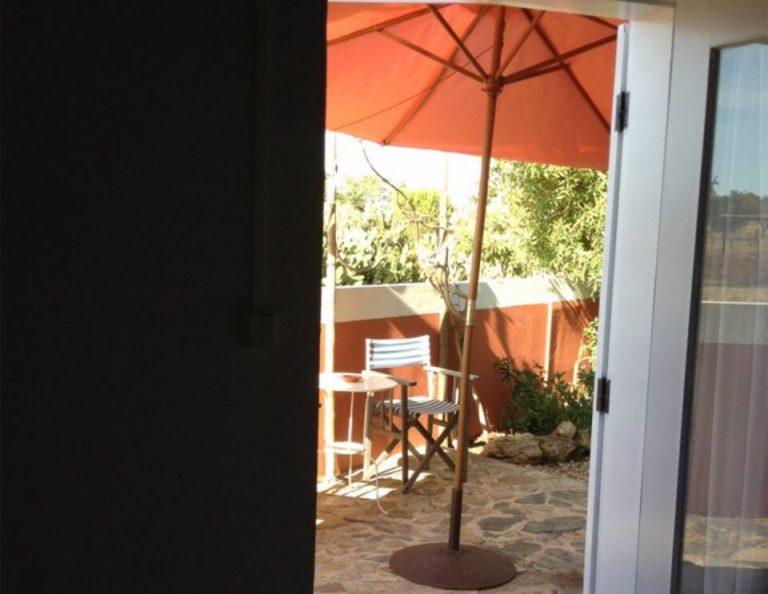 Blick durch offene Glastüren auf eine Terrasse mit einem orangefarbenen Sonnenschirm, einem gestreiften Stuhl, Steinboden und Grünpflanzen unter sonnigen Bedingungen.